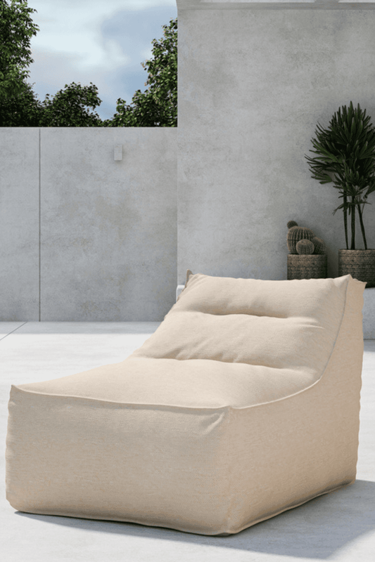 Beige outdoor lounge chair with a soft, cushioned design, placed on a modern concrete patio against a minimalistic gray wall, complemented by tall green plants in woven pots, creating a cozy and stylish outdoor relaxation space. | PicSEO 1773144872841