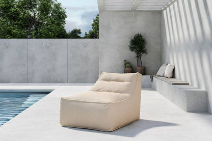 A modern outdoor poolside lounge area featuring a beige cushioned bean bag chair on a smooth concrete deck, complemented by built-in seating with pillows, minimalist concrete walls, lush green plants in woven baskets, and natural sunlight creating striped shadows from a pergola overhead. | PicSEO 1773144901706