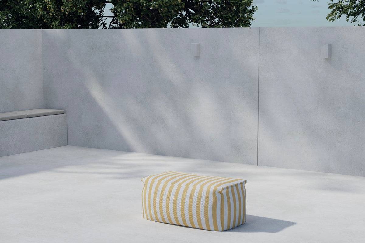 Minimalist outdoor patio setting featuring a light gray concrete floor and walls, with built-in bench seating on the left and a yellow-and-white striped rectangular pouf placed centrally, under soft natural sunlight and shaded by nearby tree foliage. | PicSEO 1773145525834