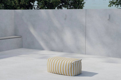 Minimalist outdoor patio setting featuring a light gray concrete floor and walls, with built-in bench seating on the left and a yellow-and-white striped rectangular pouf placed centrally, under soft natural sunlight and shaded by nearby tree foliage. | PicSEO 1773145525834
