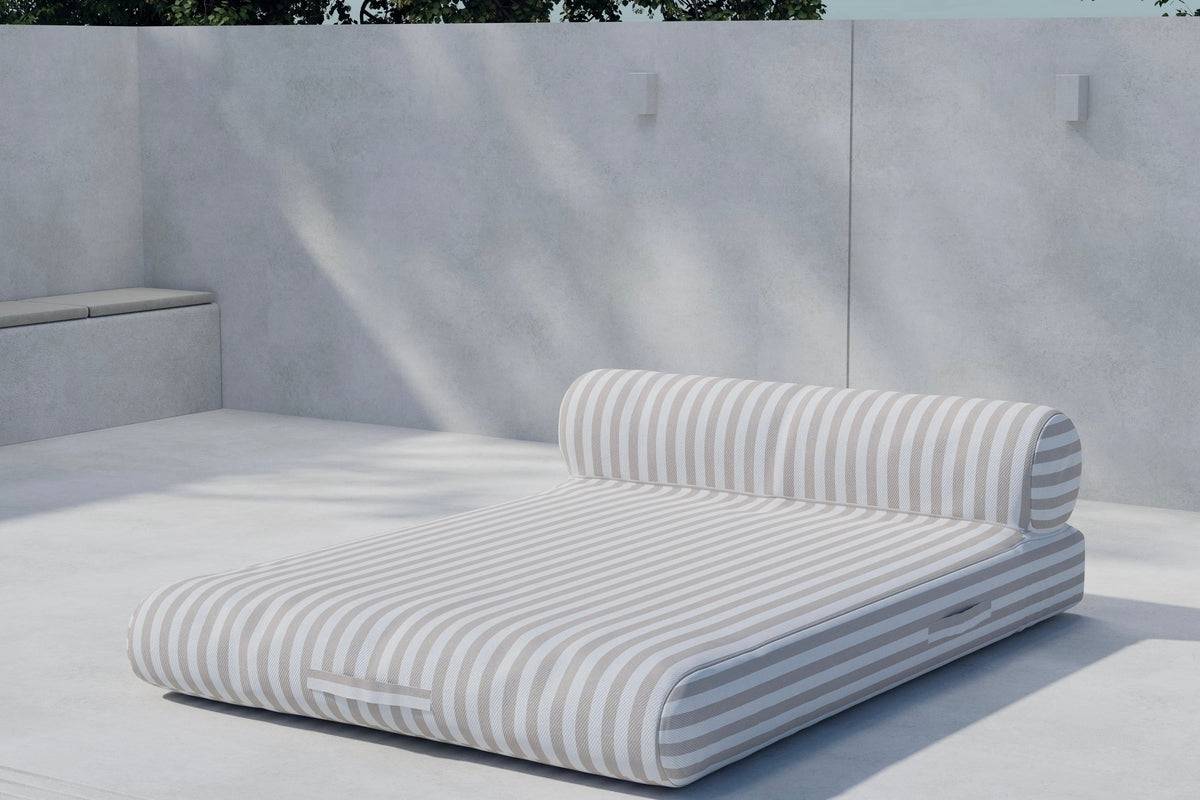Modern outdoor chaise lounge with beige and white horizontal stripes, featuring a cushioned base and cylindrical bolster pillow, set against a minimalist concrete patio backdrop. | PicSEO 1773144985486
