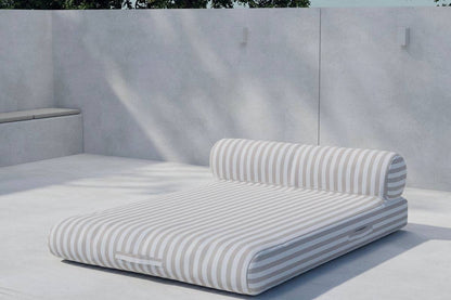 Modern outdoor chaise lounge with beige and white horizontal stripes, featuring a cushioned base and cylindrical bolster pillow, set against a minimalist concrete patio backdrop. | PicSEO 1773144985486