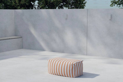 Minimalist outdoor space featuring a modern concrete patio with a built-in bench and a striped rectangular pouf in white and tan, set against a tall light gray wall with soft natural shadows and tree foliage visible above. | PicSEO 1773145232289