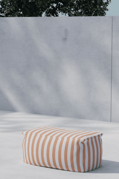 A modern outdoor setting featuring a rectangular pouf with bold orange and white stripes, placed on a smooth concrete surface against a minimalist gray concrete wall with subtle shadows and a hint of greenery above. | PicSEO 1773145199421