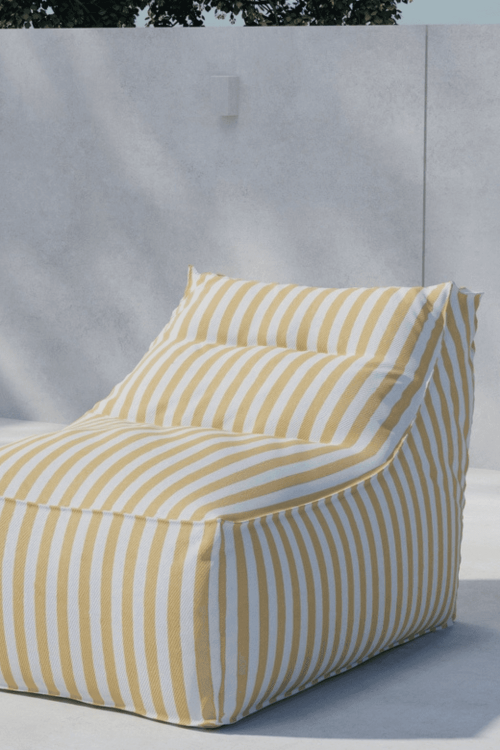 Comfortable outdoor bean bag chair with yellow and white vertical stripes, positioned against a minimalist concrete wall in a sunlit patio setting. | PicSEO 1773145541543